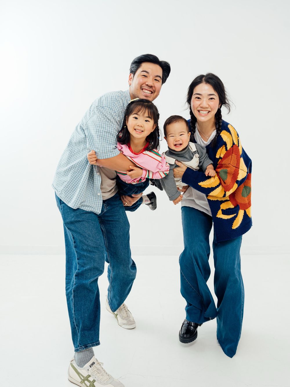 Happy Asian family of four posing together, parents holding two young children, smiling against white background.