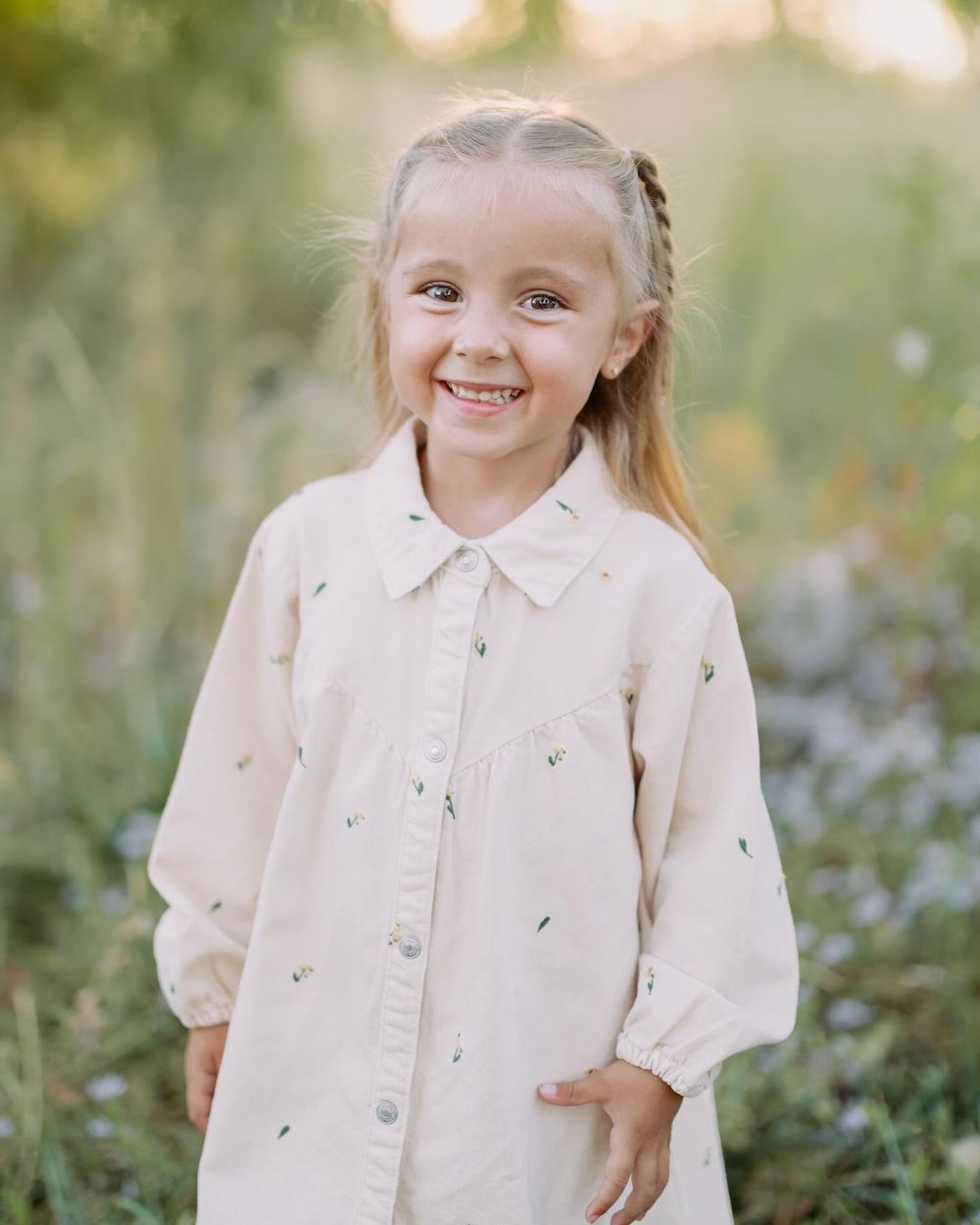 Young girl in a cream button-up dress with small floral print smiling outdoors in a sunlit field.