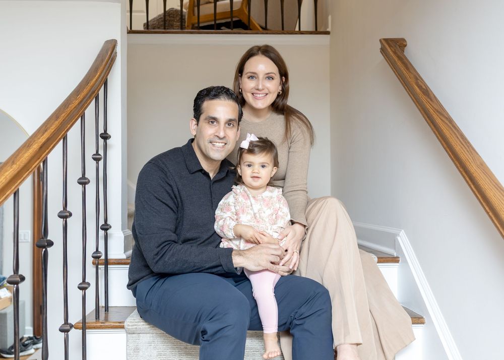 A happy family of three poses on a staircase, parents smiling with a baby girl in a floral outfit.