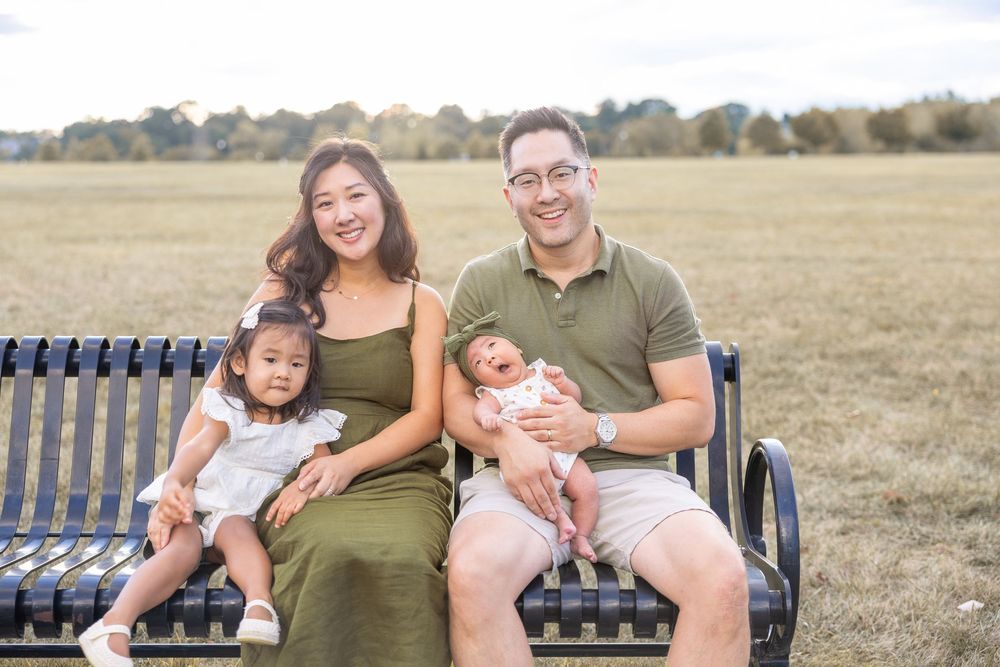 Family of four sitting on a park bench outdoors, parents with toddler and newborn baby, smiling in a sunny field.