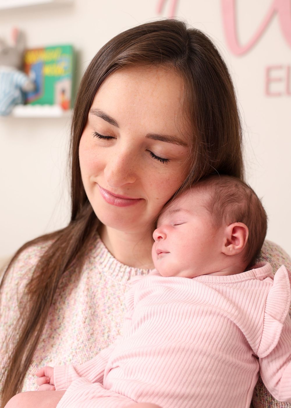A young mother with long brown hair smiles softly while cradling her sleeping newborn baby dressed in pink.