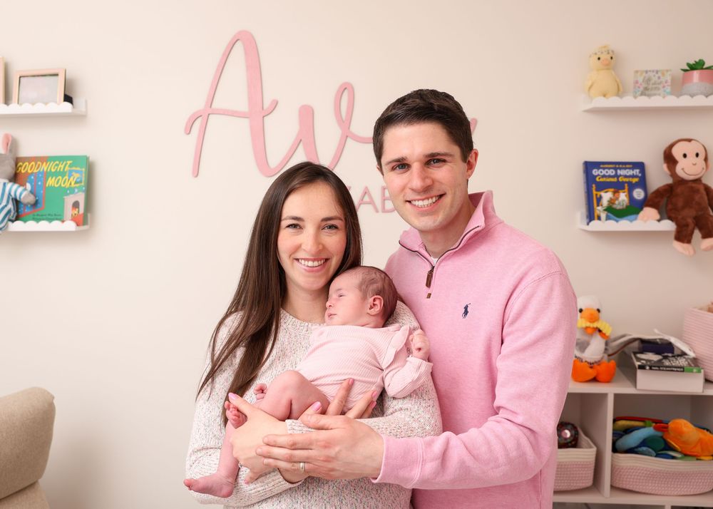 Happy couple holding their newborn baby in a pink nursery with the name Ava on the wall.