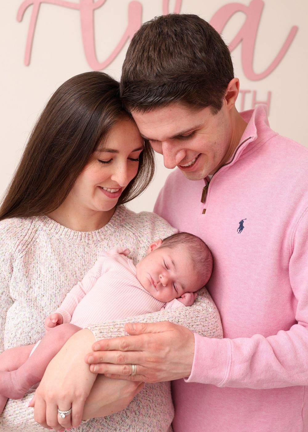 A couple in pink outfits smiles while cradling their sleeping newborn baby, posed in front of a pink name sign.