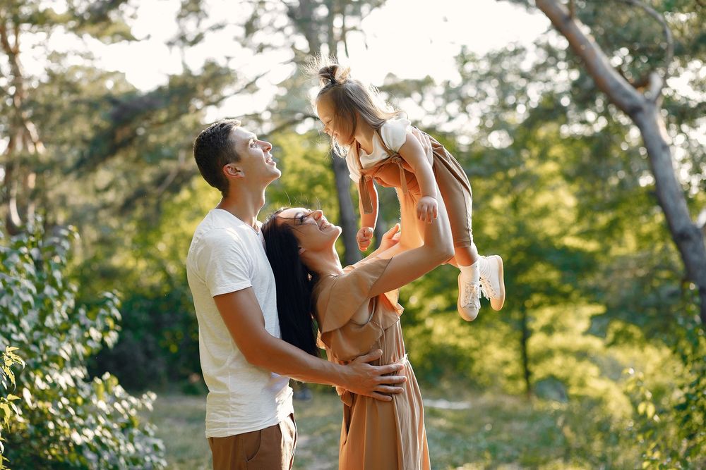 Happy family with parents lifting their young daughter in a sunny forest park during a fun outdoor adventure.