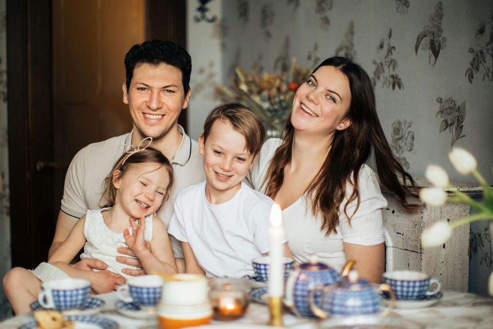 Happy family of four smiling together at a table with blue and white tea set in a cozy home setting.