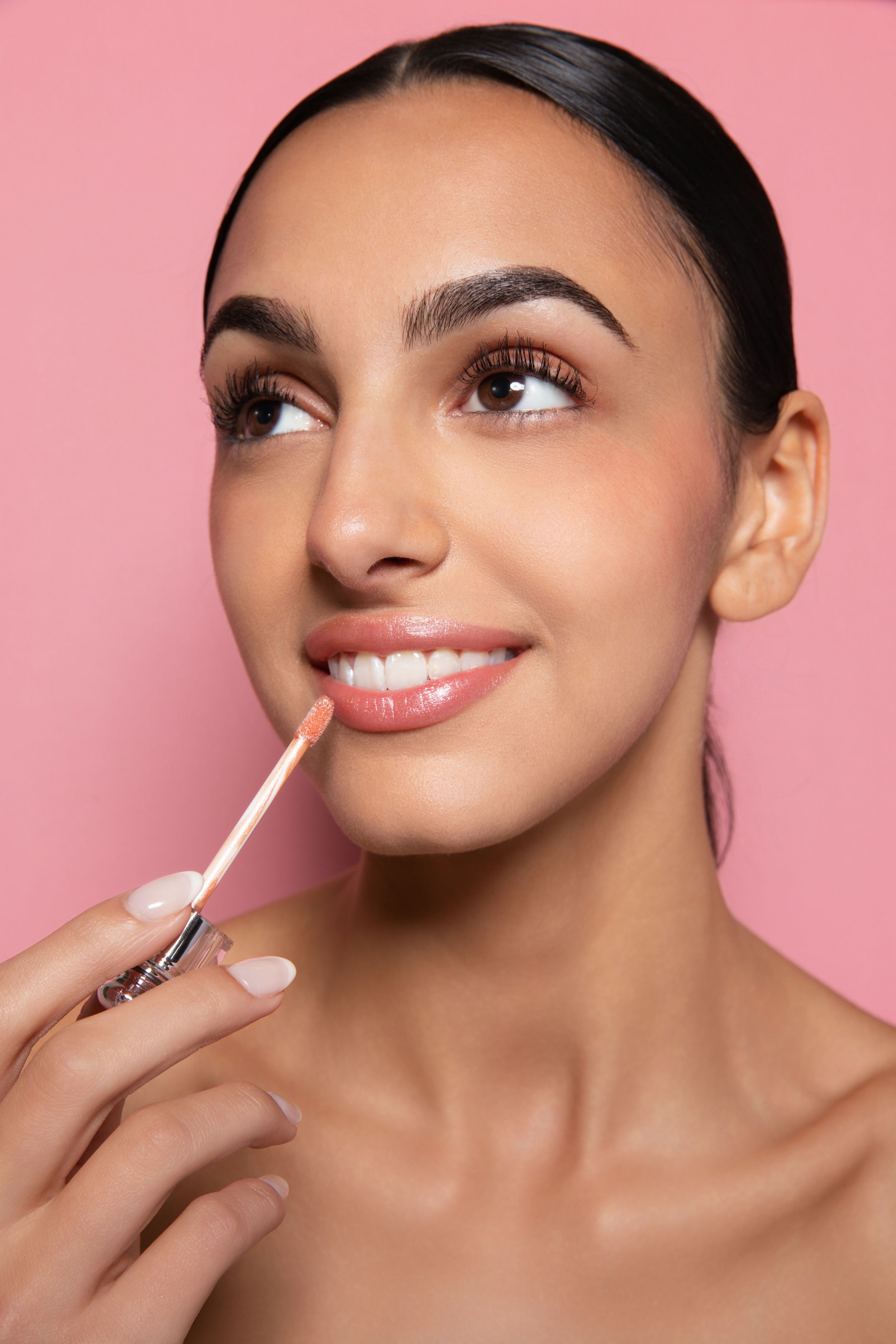 Young woman applying nude lip gloss with applicator, smiling against pink background in beauty makeup shot.