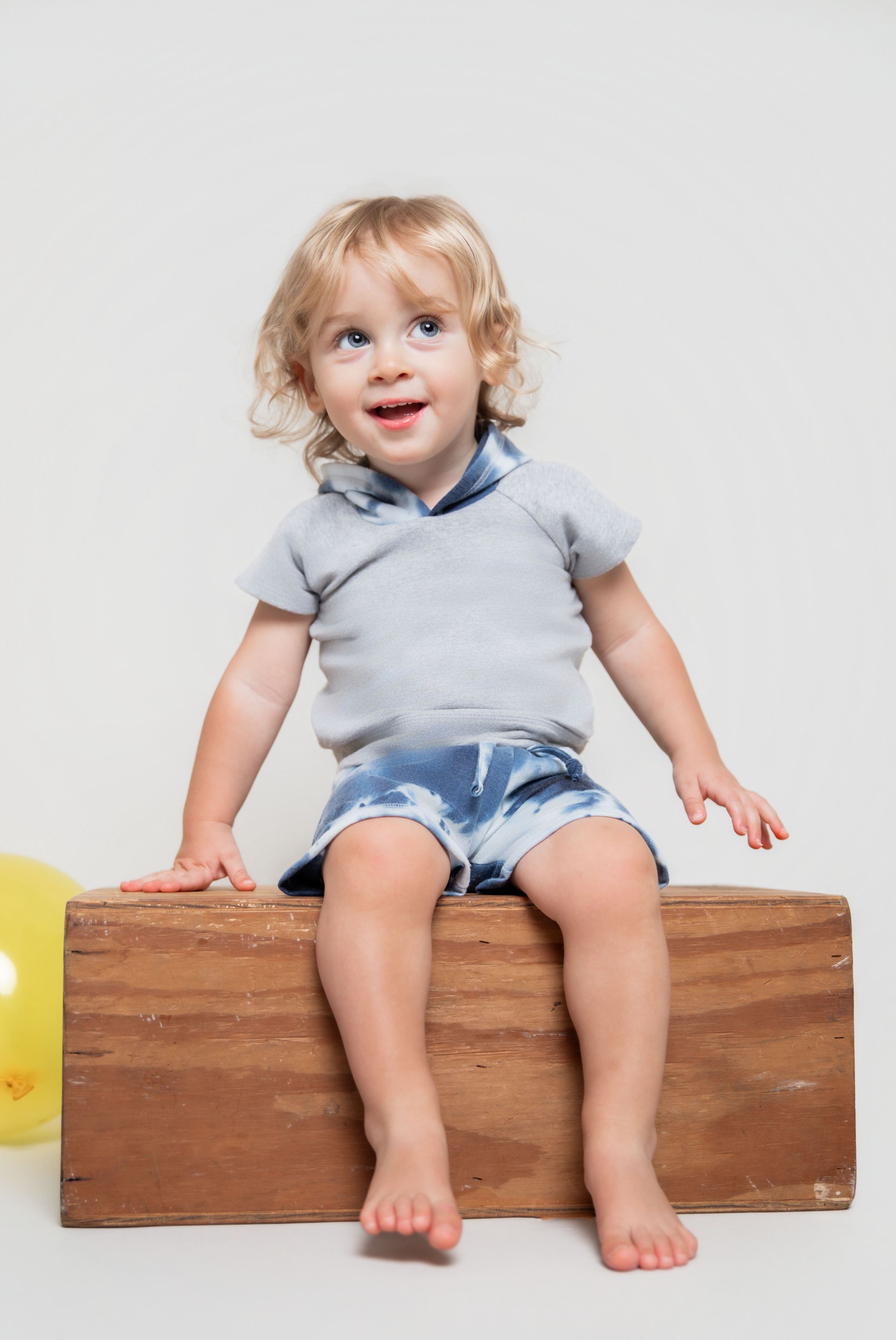 Smiling toddler with curly blonde hair sitting on wooden box wearing gray shirt and denim shorts with yellow balloon nearby.