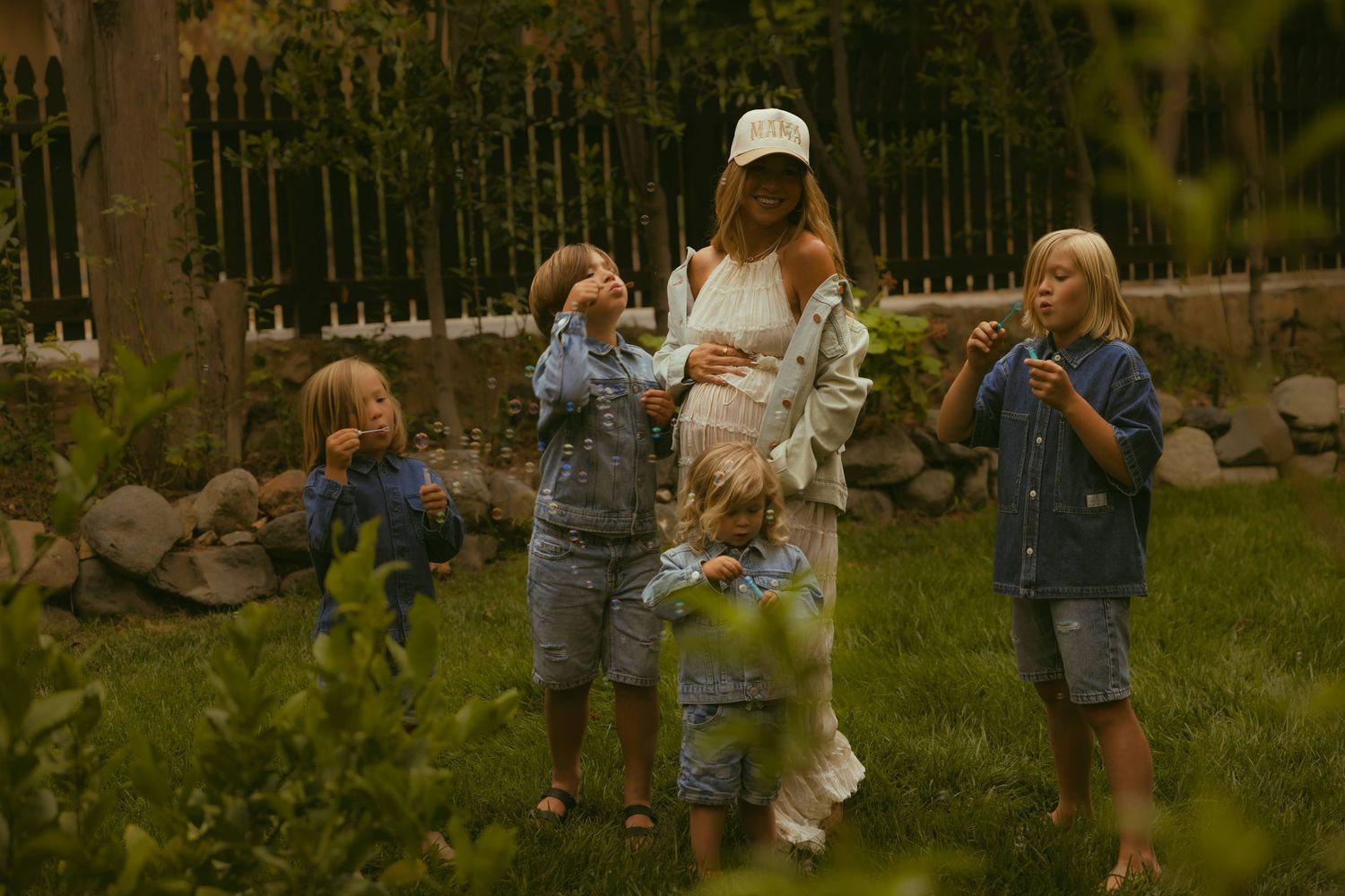 A pregnant woman surrounded by four young children playing outdoors in a lush green garden.