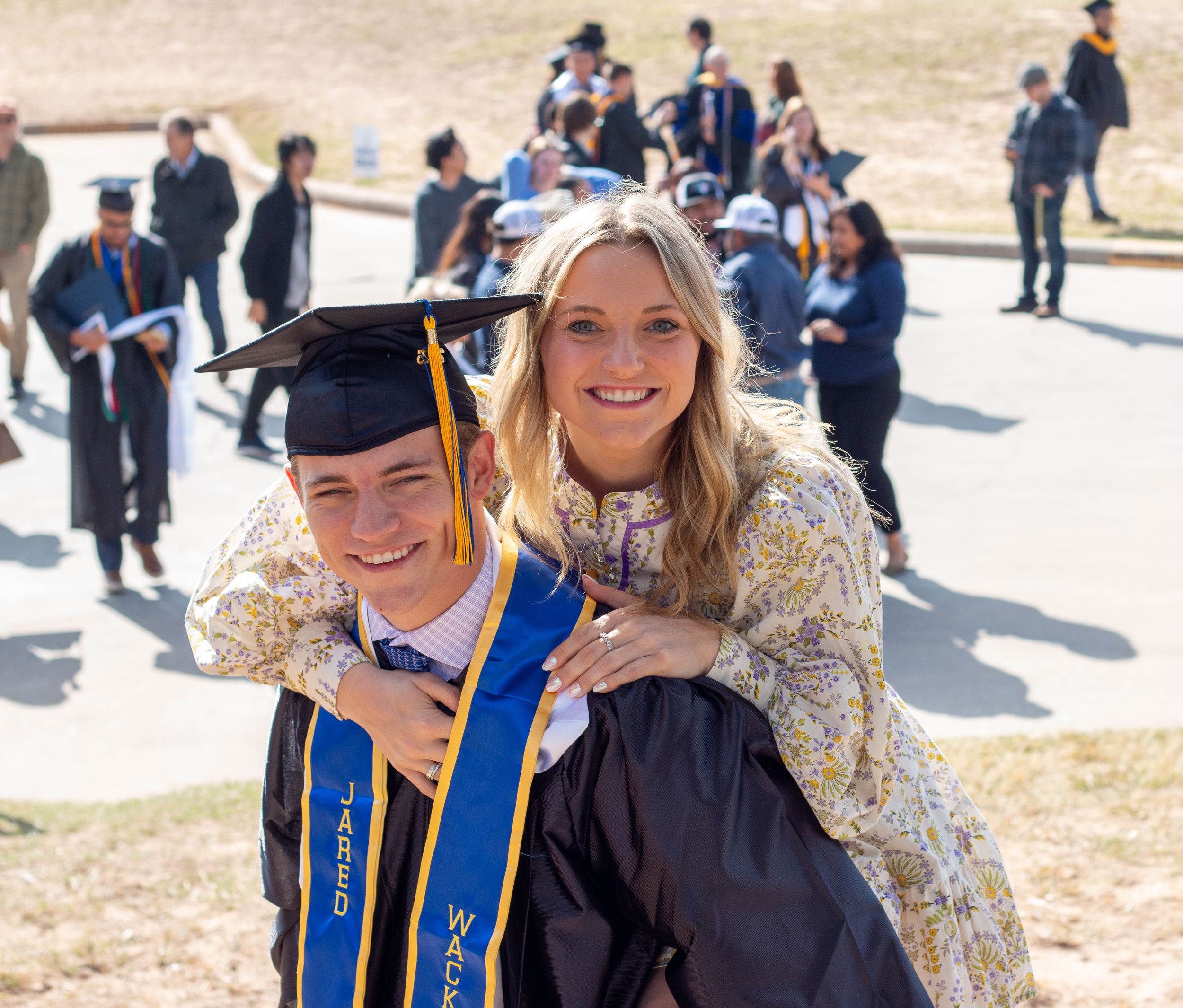 Graduate in cap and gown smiles with proud woman at outdoor college graduation ceremony.