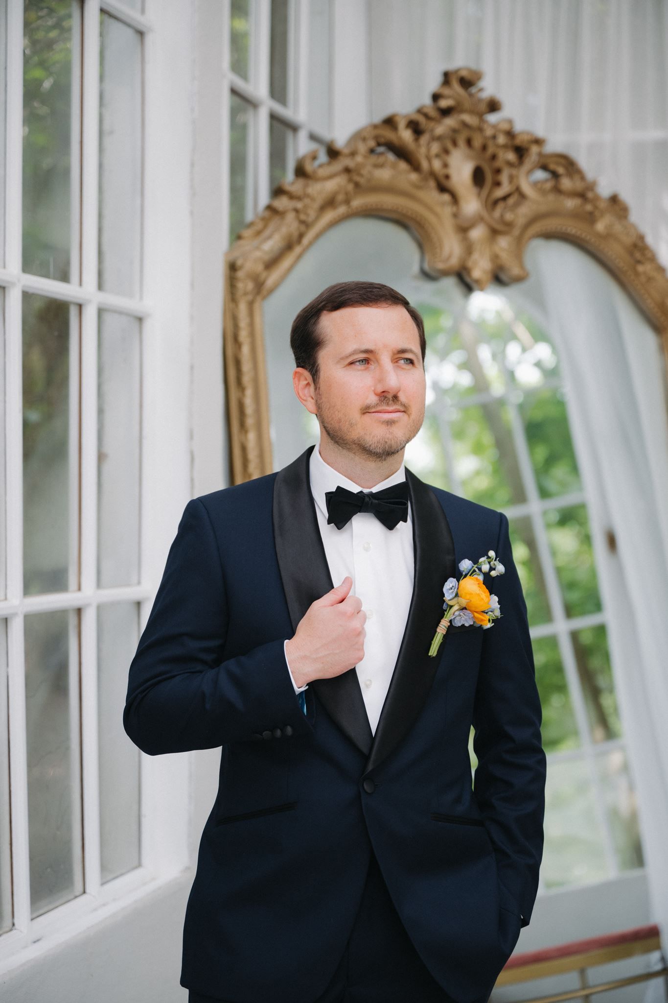 Groom in navy tuxedo with bow tie and boutonniere poses before ornate gold mirror in elegant venue.