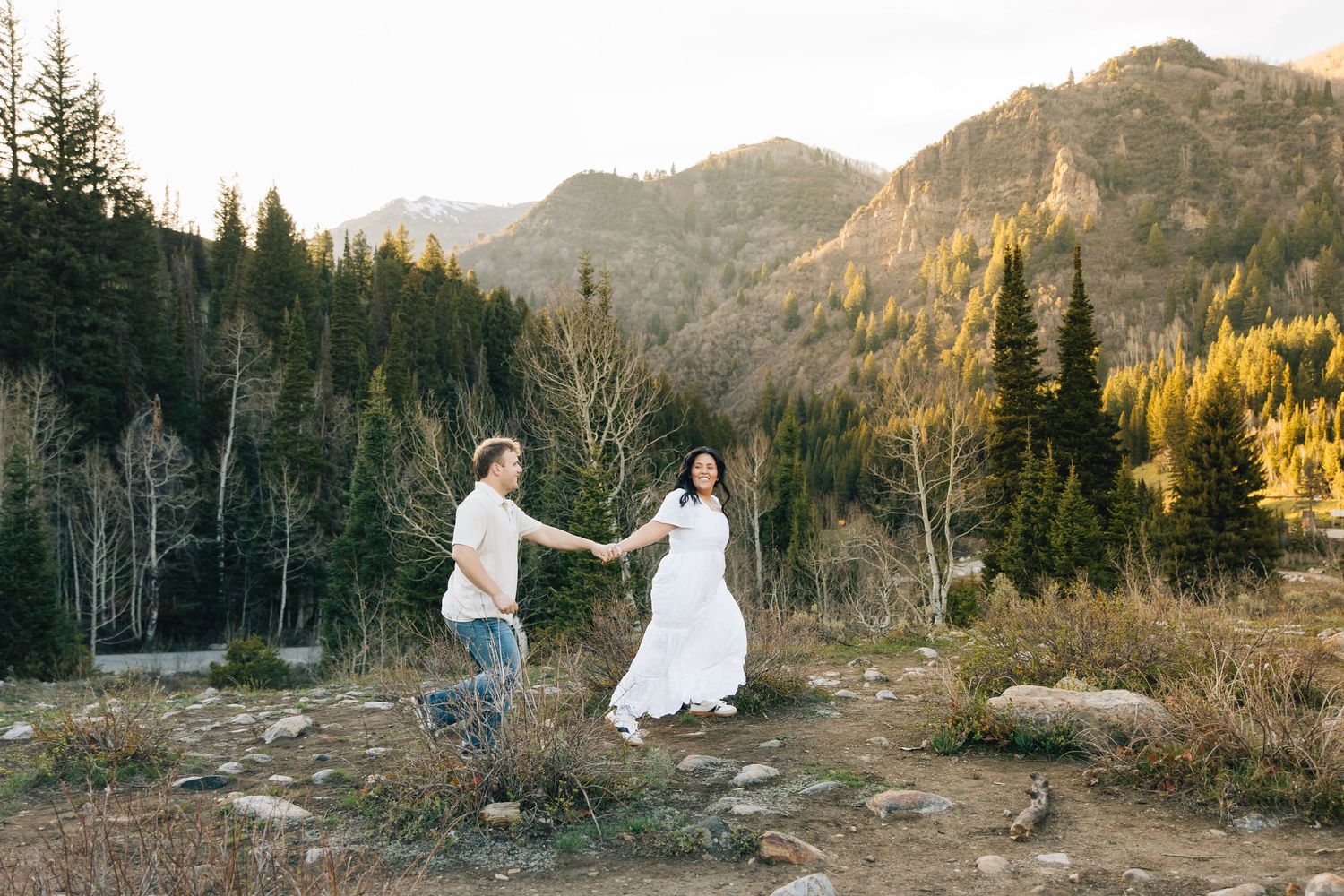 Couple holding hands in white wedding attire with mountain forest backdrop at golden hour.