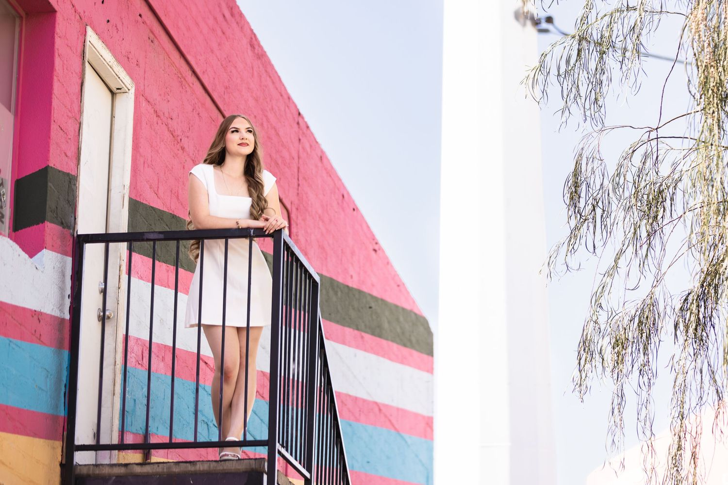Young woman in white dress stands on colorful pink mural building balcony, gazing upward on a bright day.