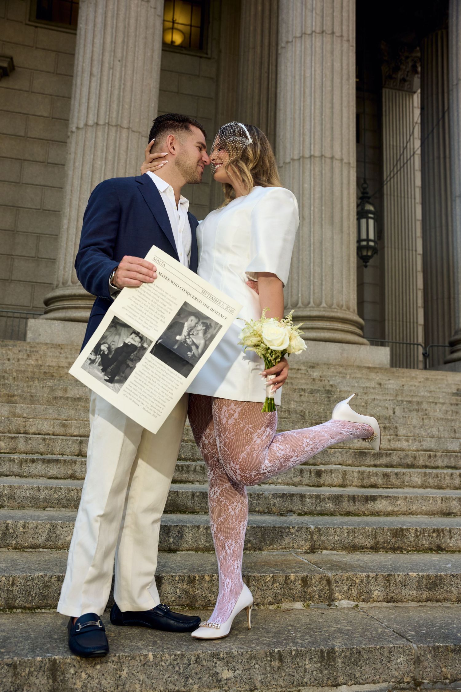 Newlywed couple sharing a romantic kiss on stone steps, bride in white mini dress with lace tights holding bouquet.