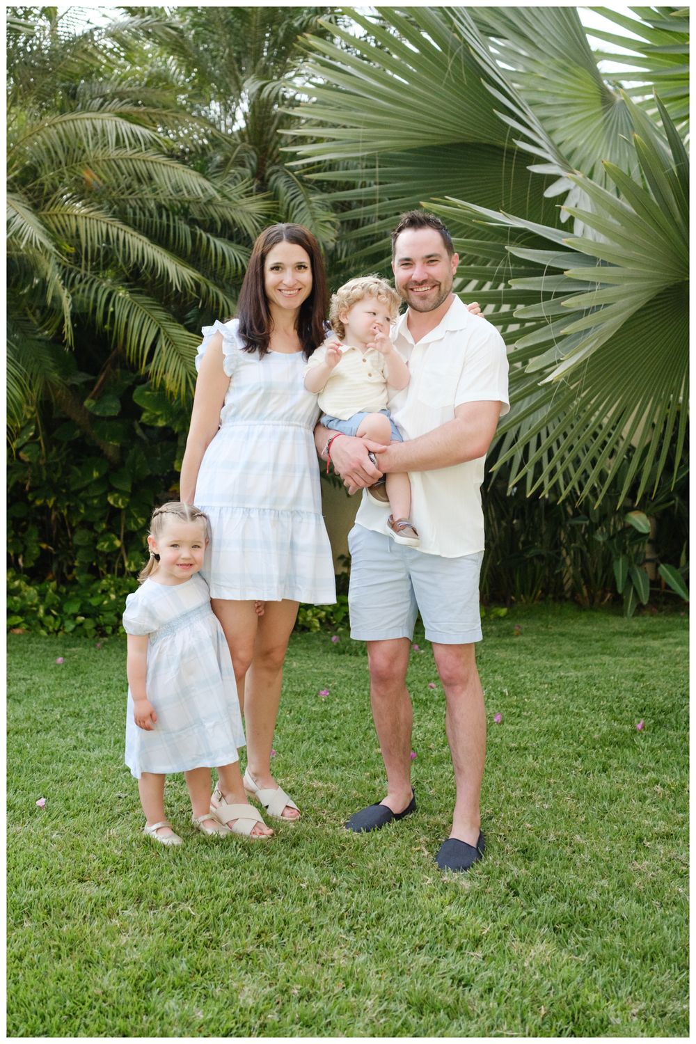 Family of four posing outdoors on green grass with tropical palm trees, wearing coordinated white and light blue outfits.
