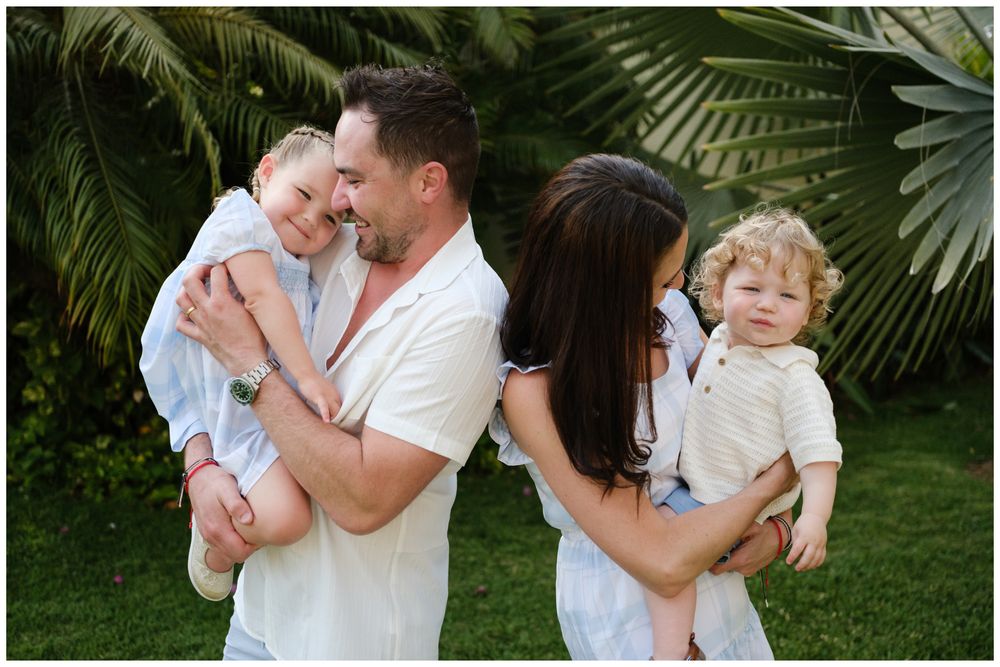 Family of four posing outdoors with tropical palms, parents holding two young children in white outfits.