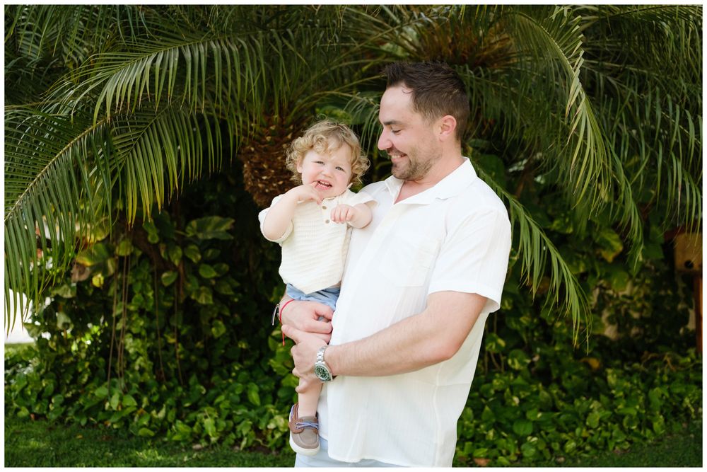 Father holding toddler child outdoors near tropical palm trees, sharing a joyful moment in a lush green garden setting.