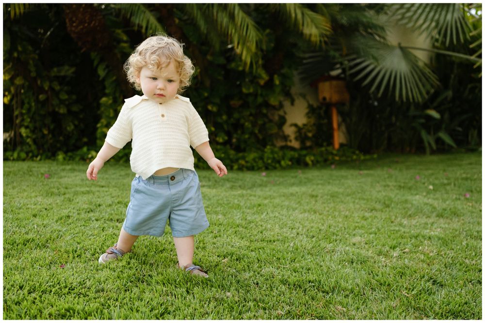 Toddler with curly hair wearing cream top and blue shorts walking on green grass with tropical plants behind.