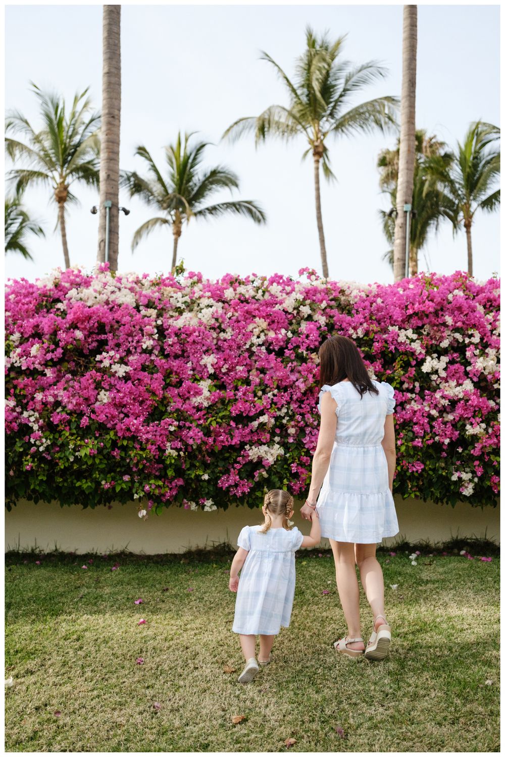 Mother and daughter in white dresses holding hands before vibrant pink bougainvillea hedge with tropical palm trees.