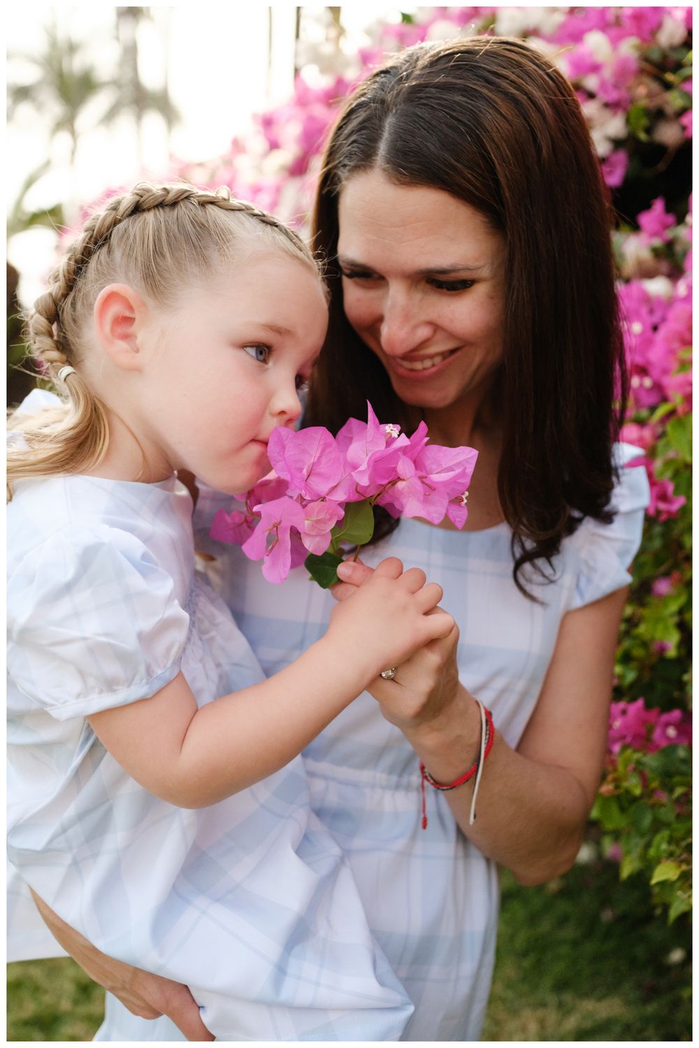 A smiling mother holds pink bougainvillea flowers for her young blonde daughter to smell outdoors.