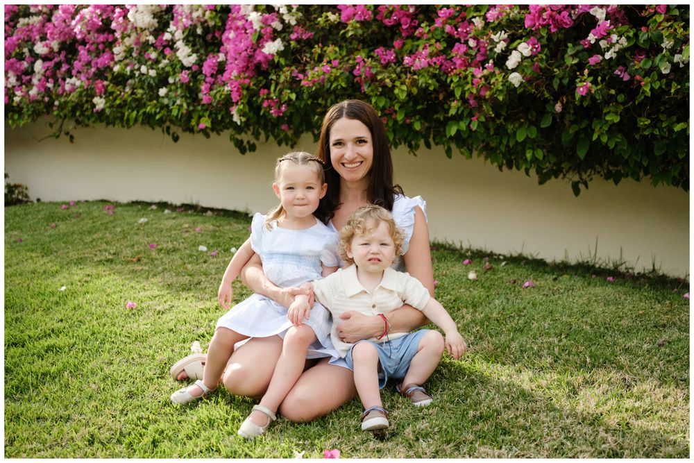 A smiling mother sits on green grass with two young children in front of a blooming pink floral wall.