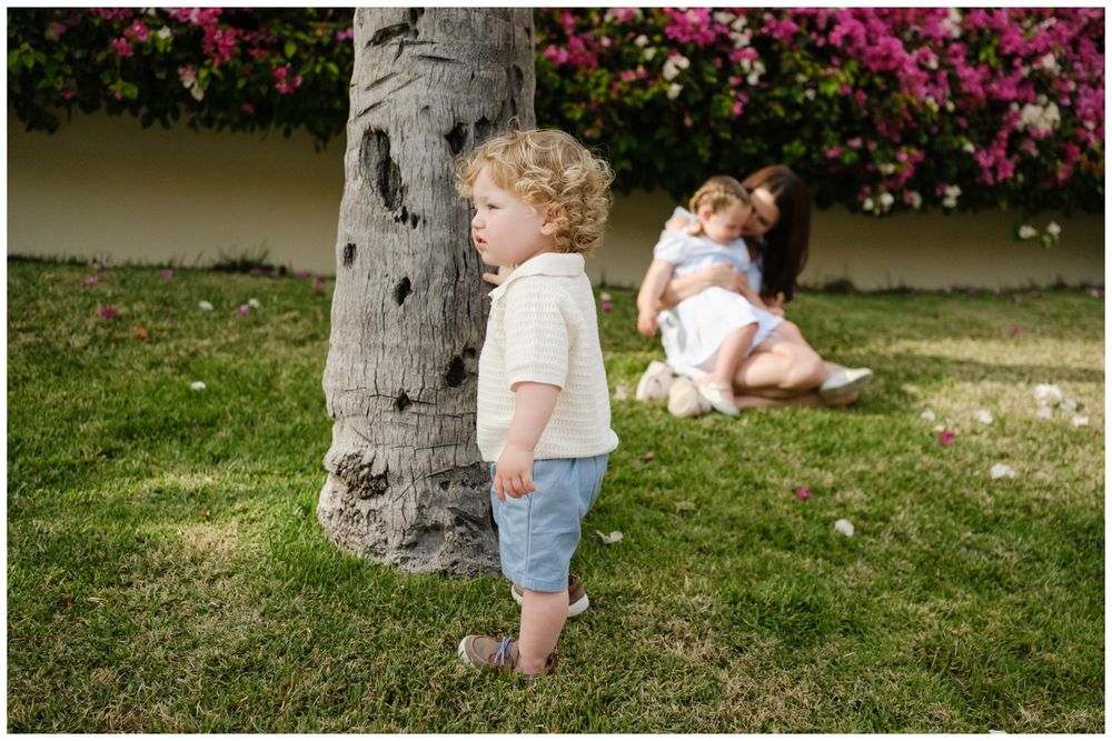 Toddler with curly hair stands by palm tree while mother sits on grass with baby near pink flowers.