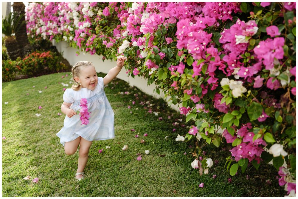 Toddler girl in white dress reaching for pink bougainvillea flowers on a sunny green lawn.