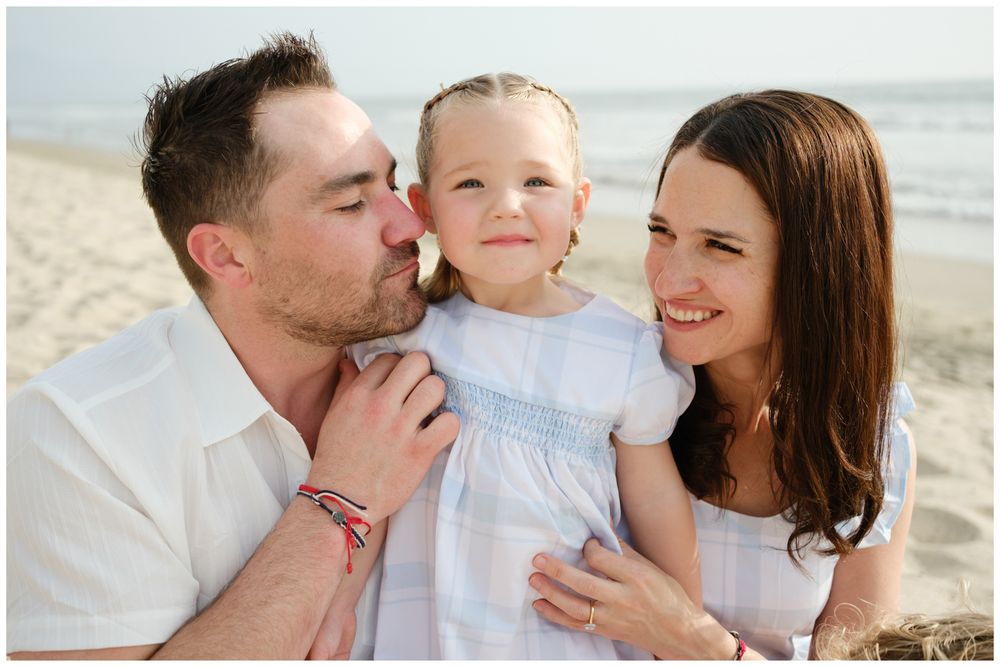 A happy family of three poses on a beach, with parents kissing and hugging their young blonde daughter in a white dress.