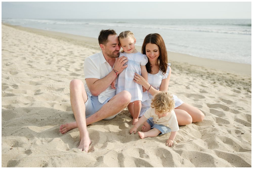 Family of four sitting on sandy beach, parents smiling at toddler in white dress, sunny day.