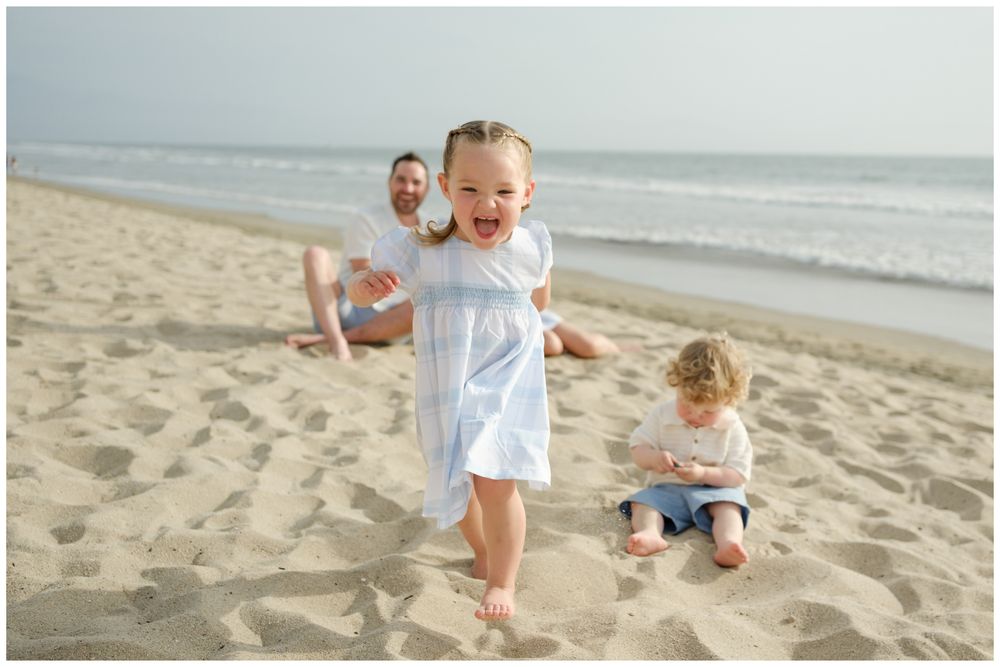 A joyful toddler girl in a blue dress runs on a sandy beach while family relaxes behind her near the ocean.