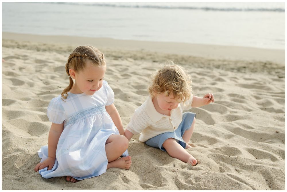 Two young children sitting on sandy beach, girl in blue dress and curly-haired toddler playing together.