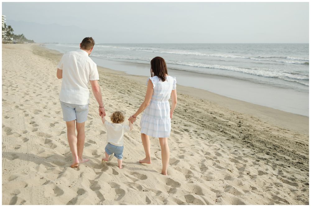 Family of three walking hand-in-hand on a sandy beach, parents swinging toddler between them near ocean waves.
