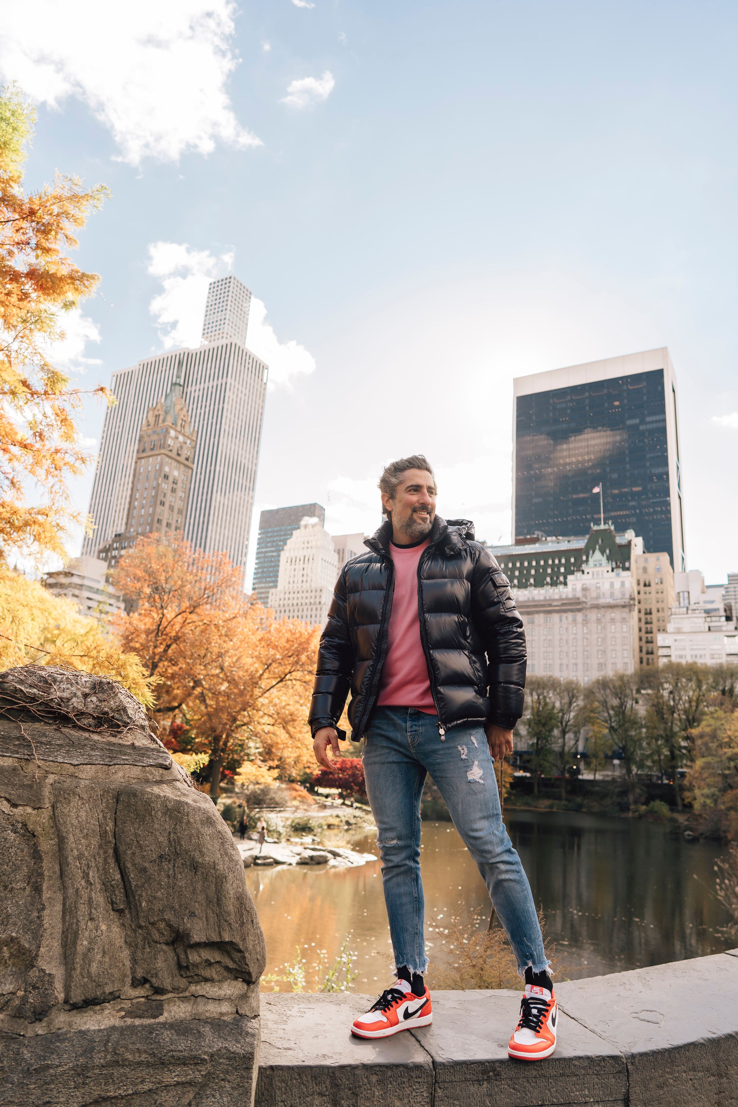 Man in black puffer jacket and ripped jeans poses in Central Park with NYC skyline in autumn backdrop.