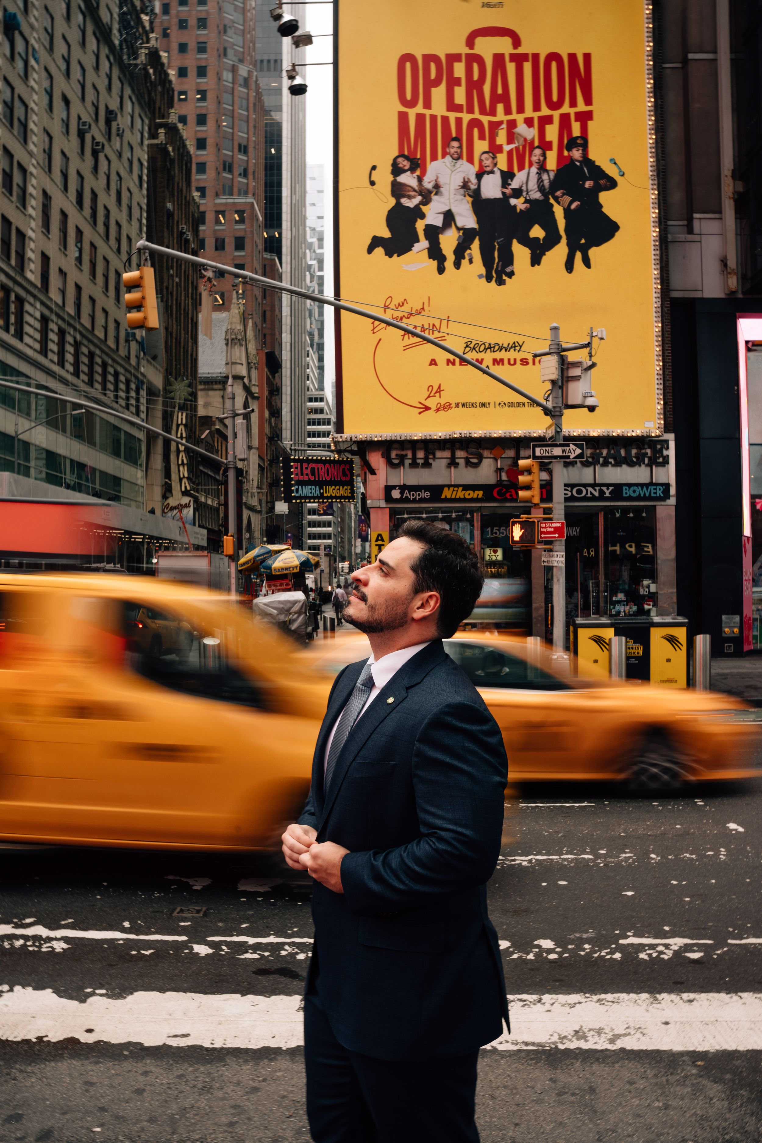 Man in suit standing in Times Square NYC with yellow taxis and Operation Mincemeat Broadway billboard.
