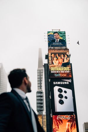 Man in suit looks up at Times Square billboards featuring Samsung, Coca-Cola, and Britbox ads against a cloudy sky.