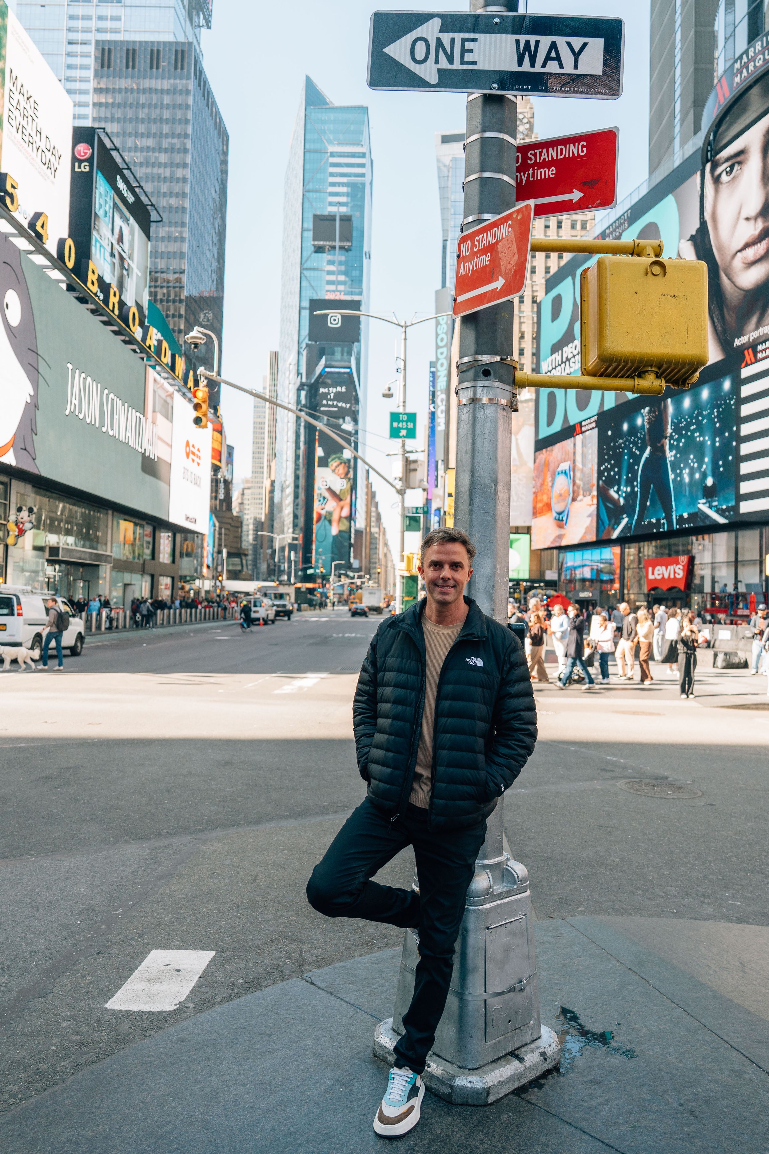 Man in black puffer jacket posing at Times Square NYC intersection with One Way sign and colorful billboards.