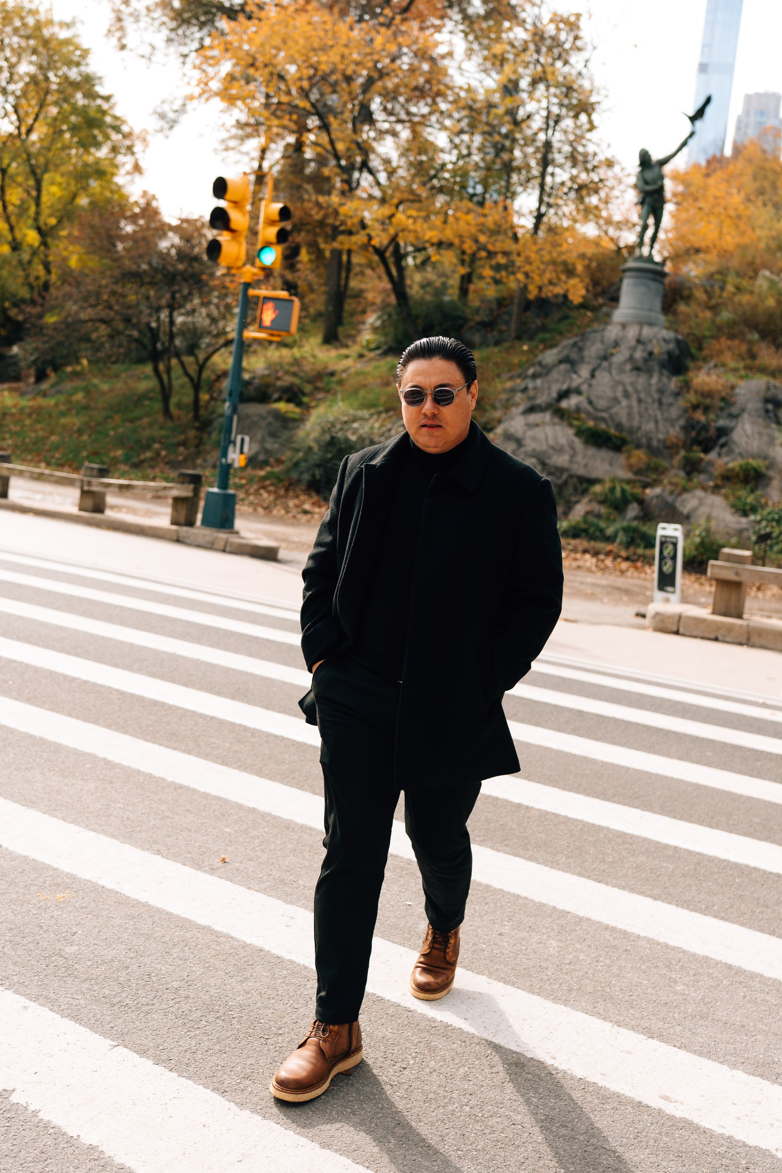 Man in black coat and sunglasses stands at crosswalk in Central Park, NYC, during autumn with colorful foliage.
