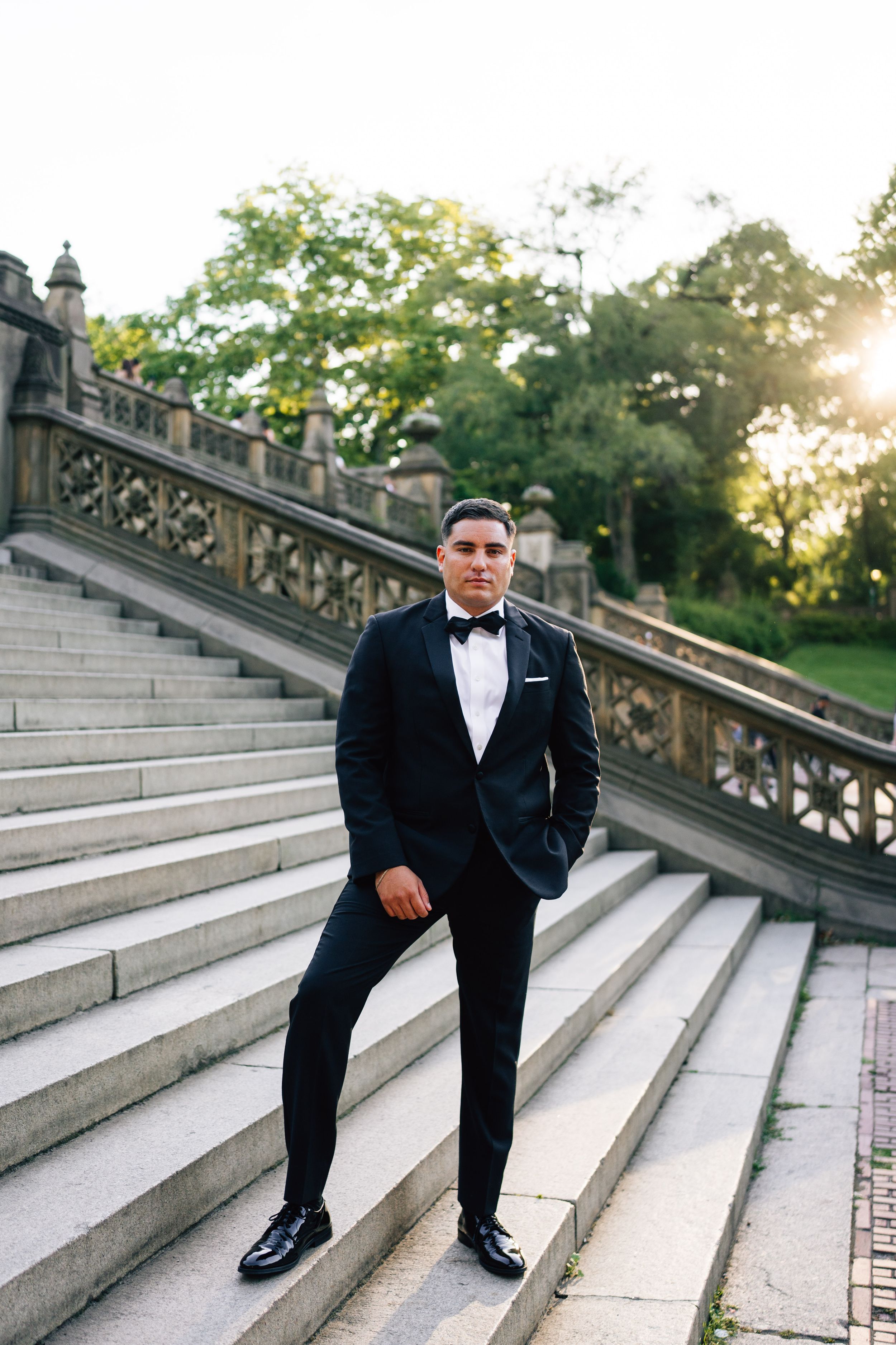 Man in black tuxedo and bow tie standing on stone steps in a park at golden hour.
