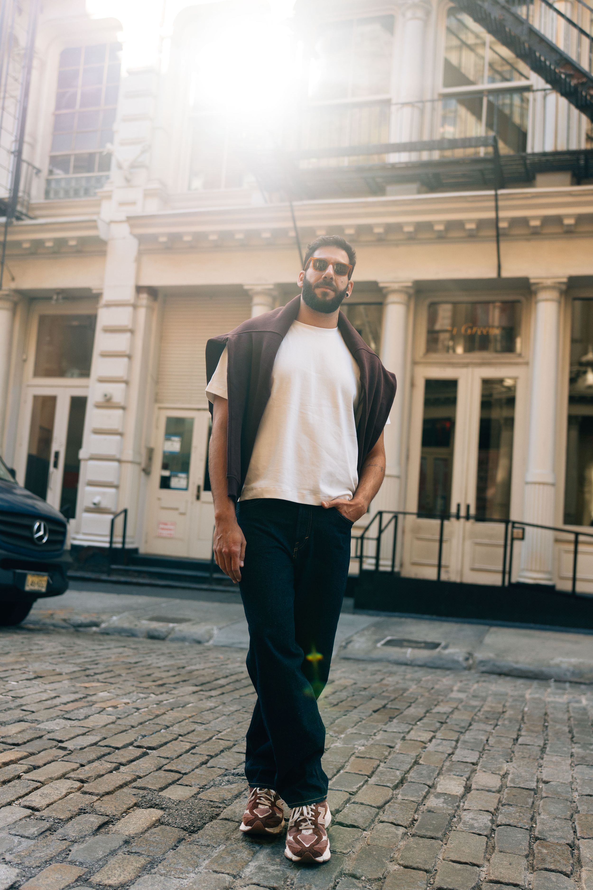 Man in white t-shirt, dark pants, and sneakers standing on cobblestone street in front of a classic building.