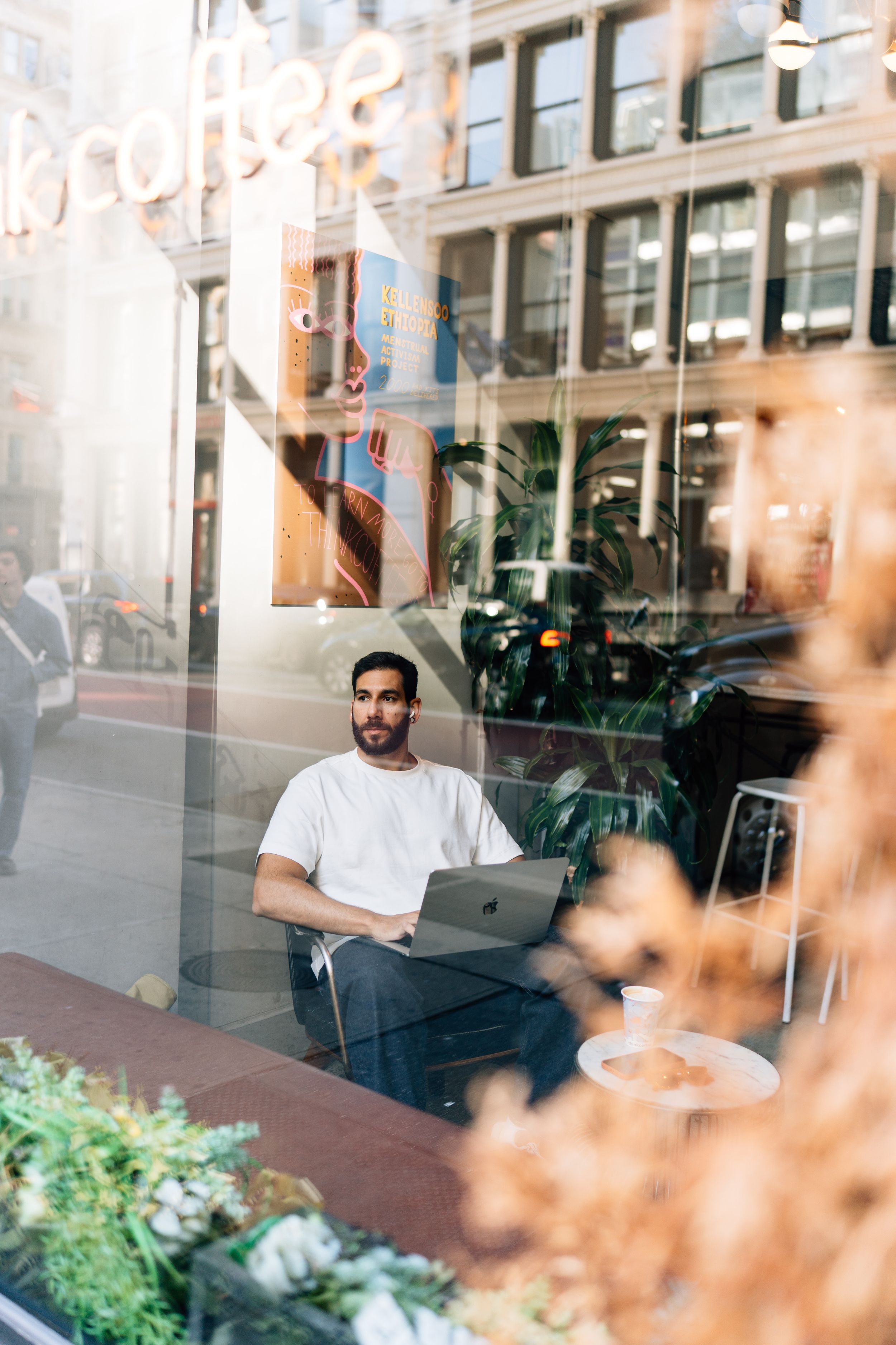 Man working on laptop in a cozy coffee shop window seat, with city reflections visible through the glass.
