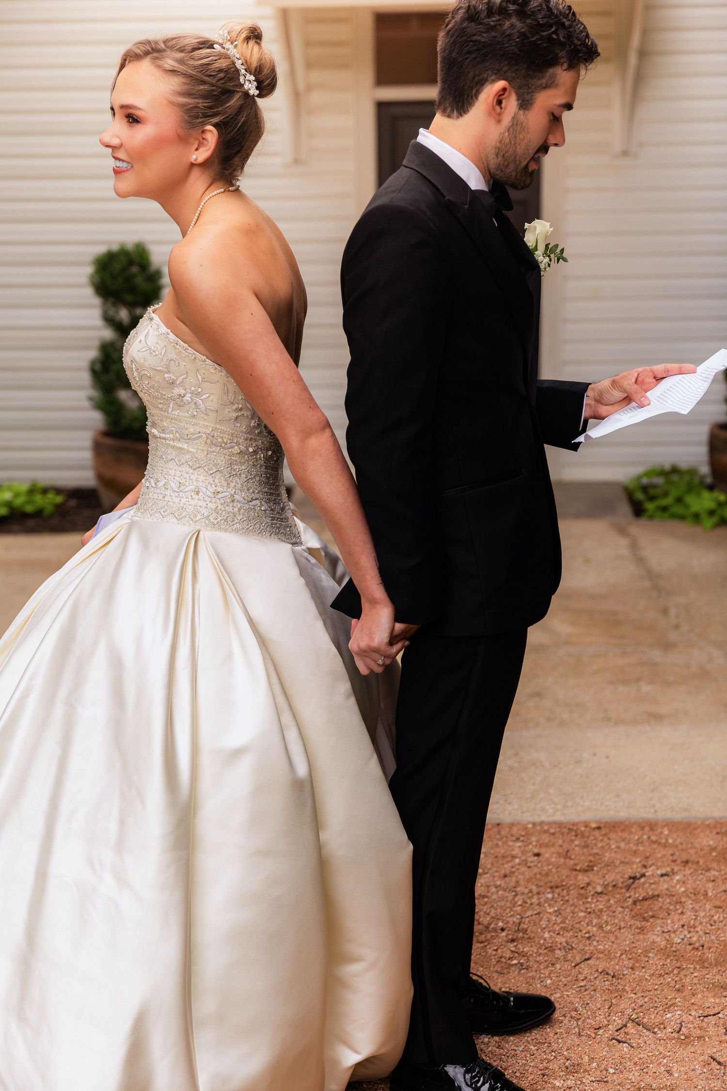 Bride in strapless gown reads a note while groom in black tuxedo stands back-to-back with her outdoors.