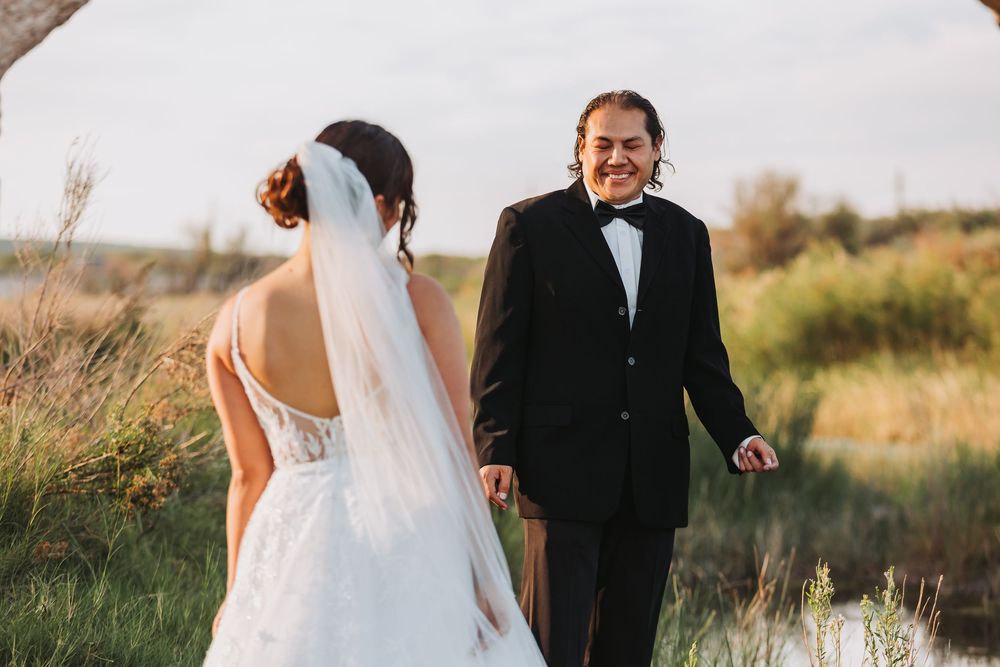 A couple in wedding attire walks through a sunlit meadow with tall grass during golden hour.
