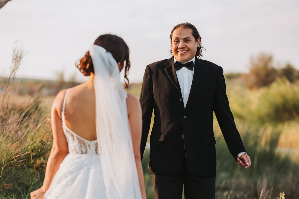 A joyful wedding couple stands in a golden field during sunset with the bride wearing a flowing veil and white dress.