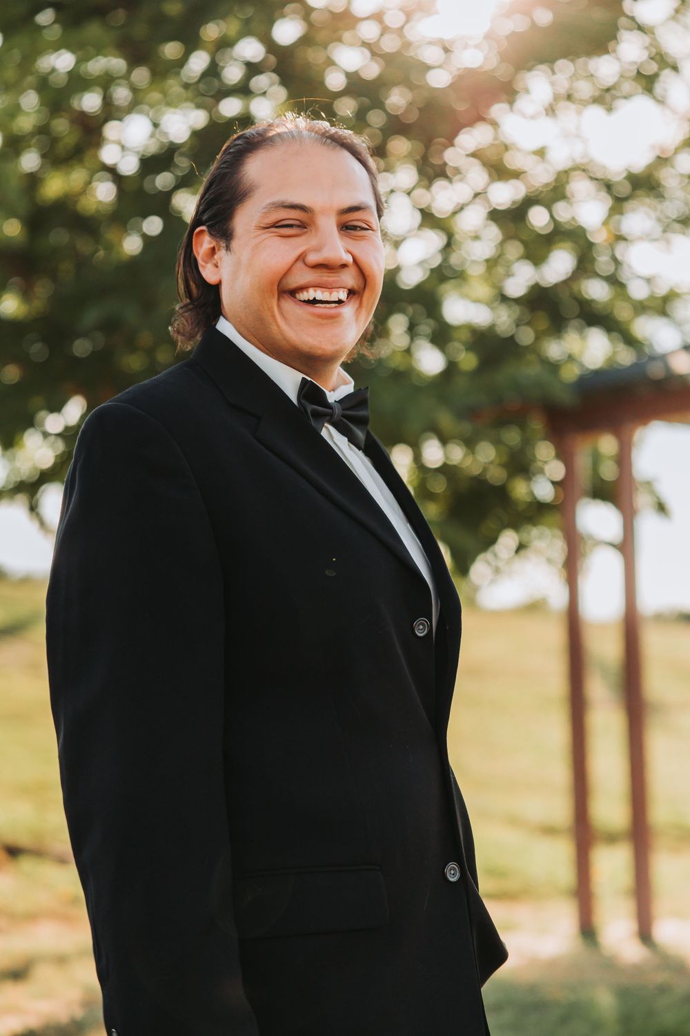 A groom in a black tuxedo smiles joyfully during an outdoor wedding photoshoot at golden hour.