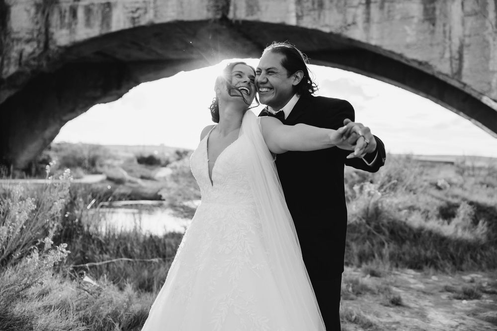 A romantic black and white wedding photo shows a couple dancing beneath a stone bridge in a natural outdoor setting.