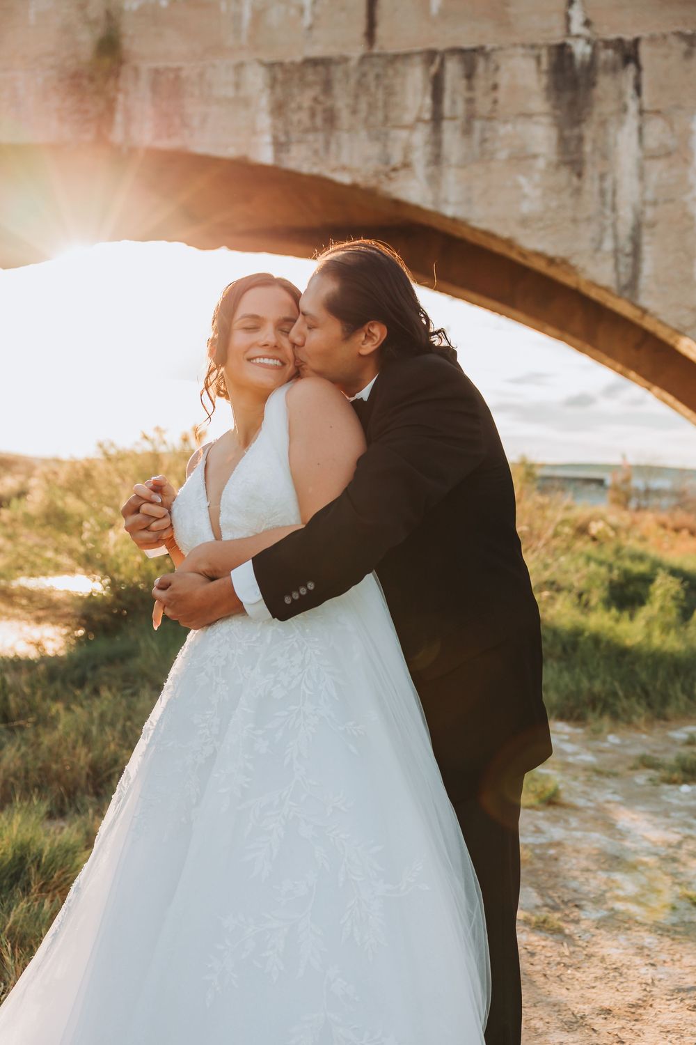 A romantic wedding couple embraces under a stone archway during golden hour sunset.