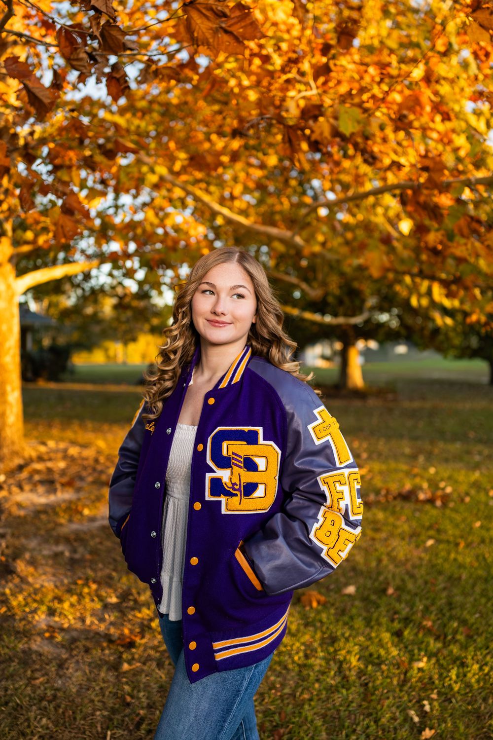 A navy blue varsity jacket with gold lettering is displayed in a fall setting with vibrant orange foliage.