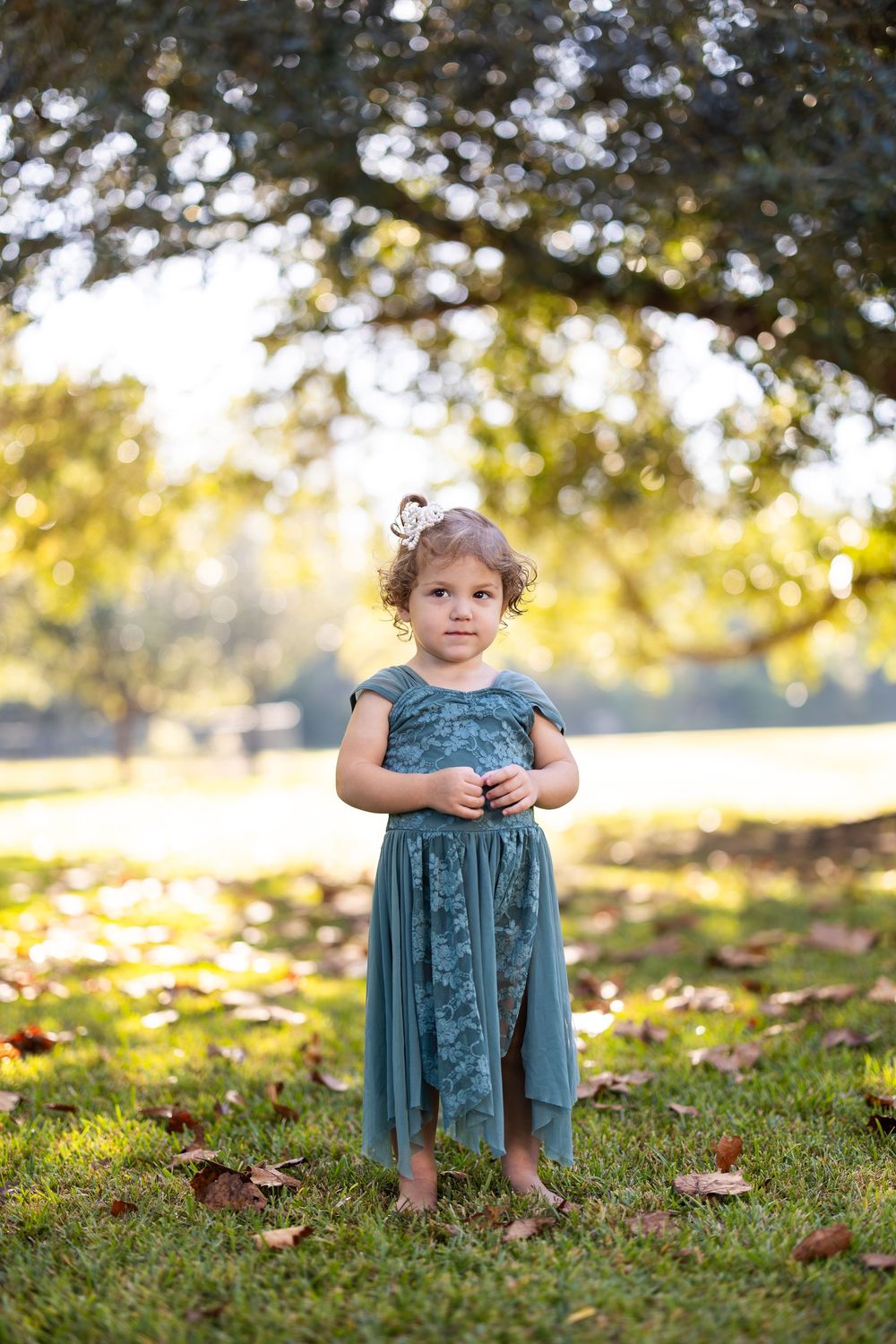 A small child in a teal dress stands on grass with fallen autumn leaves.