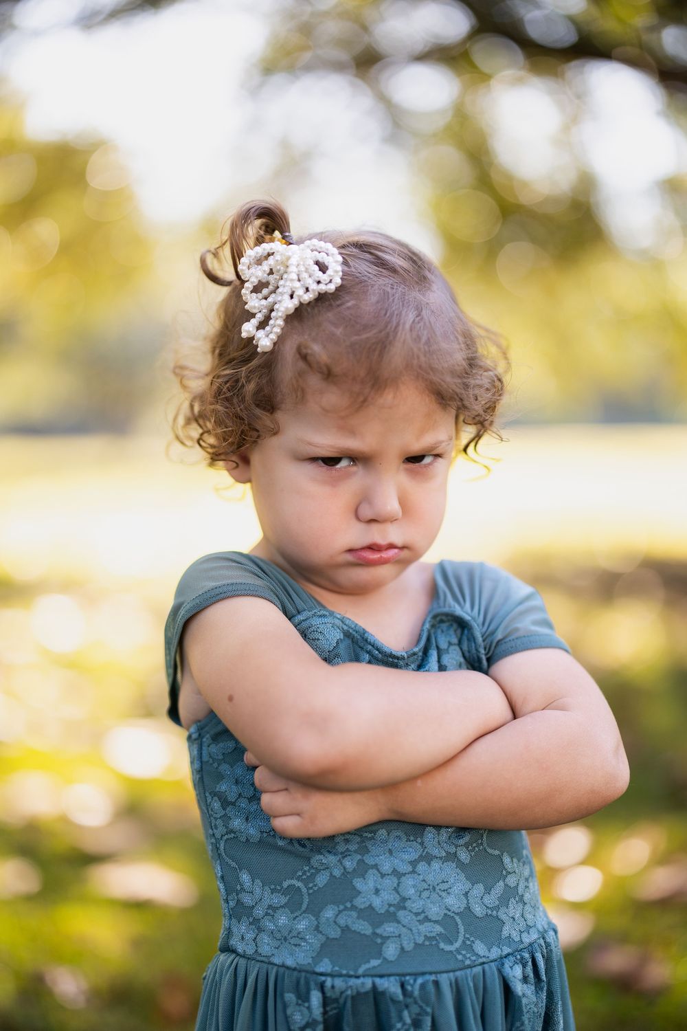 A small child in a teal dress shows an unhappy expression outdoors.