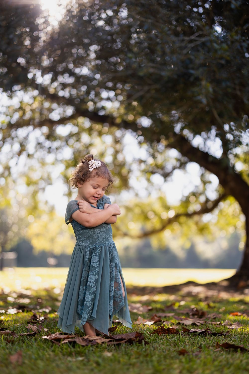 A laughing child in a teal dress stands under a tree during golden hour.