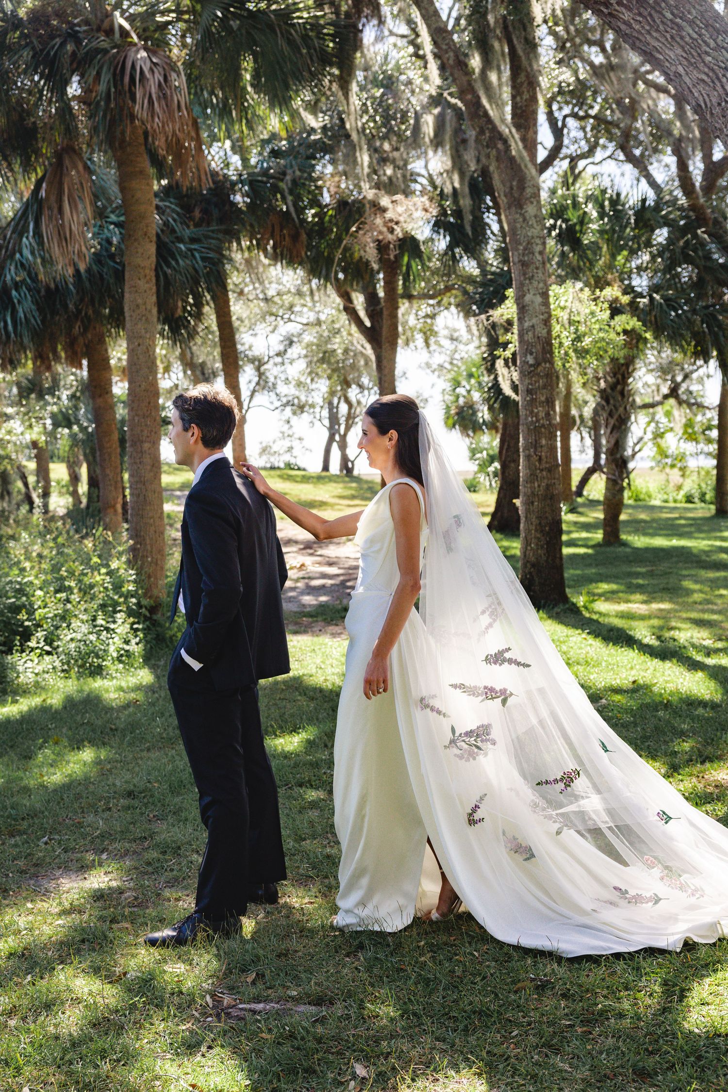 A bride in a flowing white dress with a long veil stands with her groom on a tree-lined path.